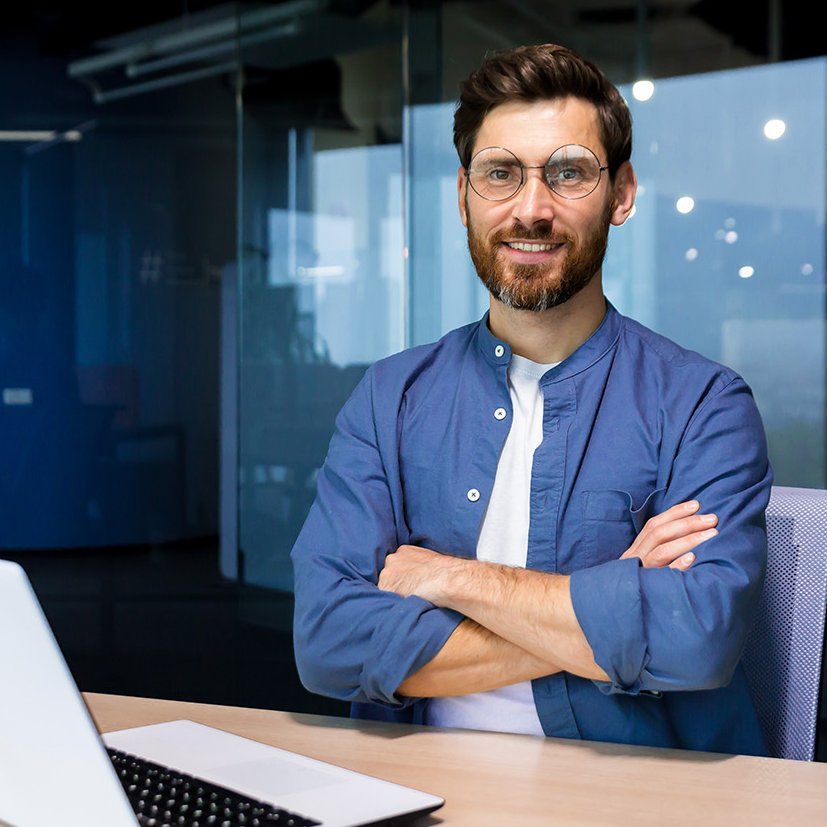 Software Entwickler gersucht Software Entwickler sitzt mit verschränkten Armen an einem Schreibtisch und lächelt in die Kamera